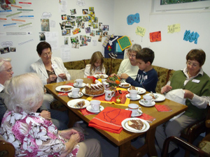 Einige Kinder und Frauen sitzen bei Kaffee und Kuchen um einen Tisch herum und arbeiten mit Nadel und Faden.