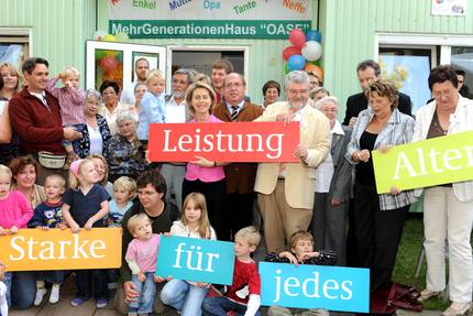Ursula von der Leyen steht mit mehreren Personen vor dem Mehrgenerationenhaus 'OASE'. Sie halten den Schriftzug 'Starke Leistung für jedes Alter' in den Händen.